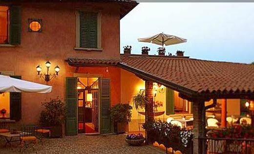 La Valletta Relais exterior view featuring lit windows, open shutters, outdoor seating under umbrellas, and flower decorations, creating a warm and inviting travel destination atmosphere.