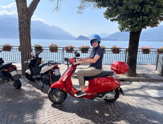 Noleggio scooter Lago di Como: uomo su Vespa rossa con casco blu, sullo sfondo panorama del lago e montagne.