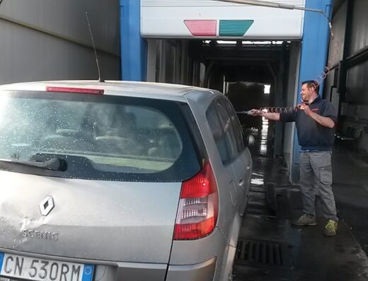 Lavaggio auto self-service: uomo lava una Renault Scenic grigia in un autolavaggio con idropulitrice ad alta pressione. Servizio di pulizia auto professionale.