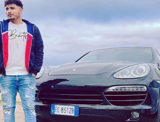 Uomo in piedi accanto a una Porsche Cayenne nera. L'auto è parcheggiata all'aperto e l'uomo indossa jeans strappati e una giacca colorata.