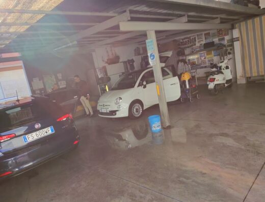 Lavaggio Auto Pastore: vista di un'area di lavaggio auto con diverse auto, tra cui una Fiat 500 bianca, un'auto nera e uno scooter bianco, in attesa di essere lavate.