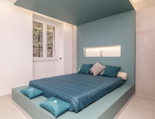 Camera da letto moderna con letto a piattaforma blu, cuscini decorativi e finestra con grata. Design minimalista e accogliente.