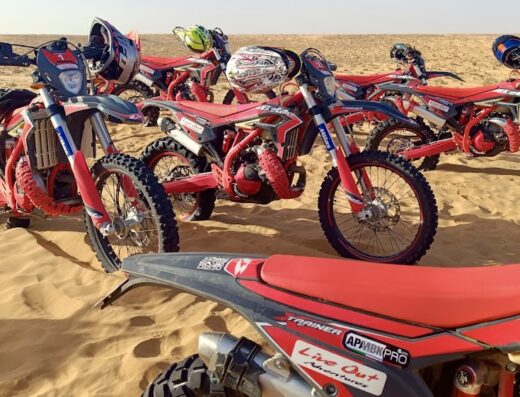 Gruppo di moto da cross rosse parcheggiate nel deserto di sabbia, pronte per un'avventura di viaggio "Live Out". Caschi da motociclista appoggiati sulle selle. Servizi di tour e marketplace per appassionati di motocross.
