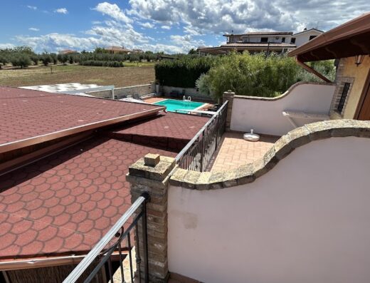 Panoramica esterna de Lo Scacciapensieri con piscina, tetto rosso e campagna circostante in una giornata di sole.