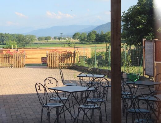 Terrazza esterna della Locanda Dl'Angel con tavoli e sedie in ferro battuto, vista panoramica sulle montagne e sulla campagna circostante. Ideale per motociclisti in viaggio, offre un'atmosfera rilassante e accogliente.