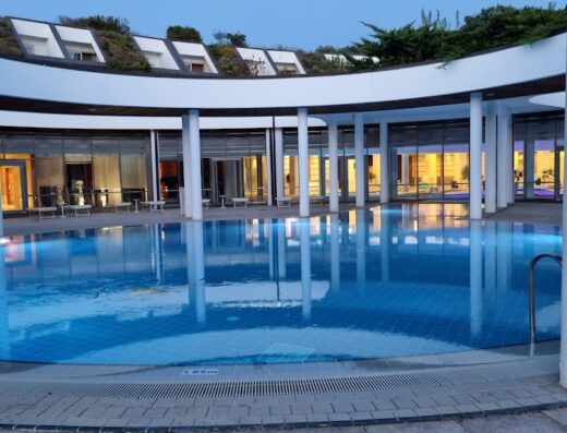 Piscina illuminata di sera in resort di lusso, design circolare con colonne bianche e riflessi sull'acqua, per vacanze e viaggi esclusivi con Lona Agenzia Viaggi.