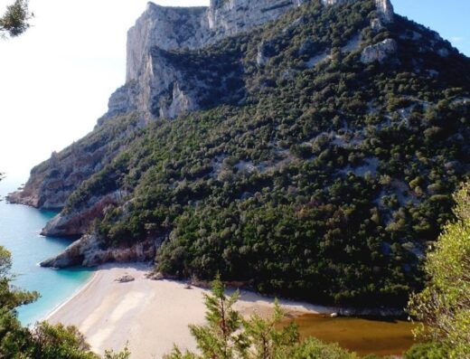 Cala di Gabbiani, Sardegna: spiaggia pittoresca con scogliera rocciosa, vegetazione lussureggiante e acque cristalline, ideale per viaggi in moto e relax.