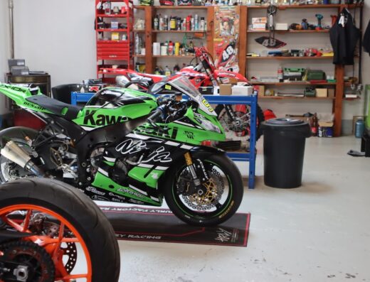 A green and black Kawasaki Ninja motorcycle is parked in a garage with other motorcycles and shelves of tools and equipment.