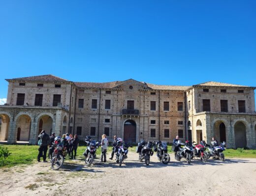 Gruppo di motociclisti LUCYRIDER in sosta con le moto davanti a un edificio storico in pietra, cielo azzurro.