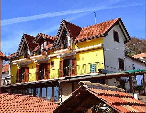 Luna D'Agerola: prospettiva di un edificio giallo a più piani con balconi e tetto rosso, situato in una zona collinare sotto un cielo azzurro.