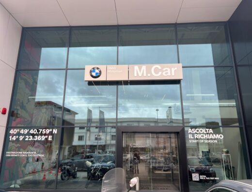 M.Car S.p.A BMW Motorrad Napoli concessionaria. Facciata in vetro con insegne BMW Motorrad e M.Car, coordinate GPS, slogan promozionali e moto esposte. Servizi e accessori per motociclisti.