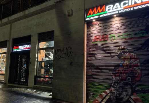 MM Racing negozio di ricambi e accessori moto e scooter, serranda con murales di motociclista, vetrine illuminate con caschi e accessori.