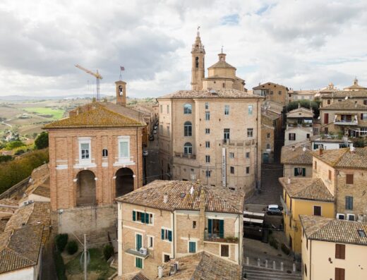 Overhead view of Corinaldo, Italy showcasing terracotta rooftops, brick buildings, a church with a dome and tower, and a construction crane against a cloudy sky.