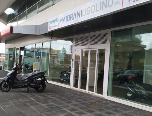 Maiorani Ugolino concessionaria moto: scooter Kymco nero parcheggiato di fronte all'ingresso, vetrine con altre moto visibili all'interno.
