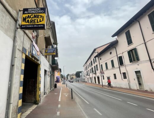 Immagine di una strada cittadina con un ciclista, insegne di "Servizio Autorizzato Magneti Marelli" e "Point Service Autofficina", che suggerisce la presenza di un gommista, meccanico e assistenza pneumatici.