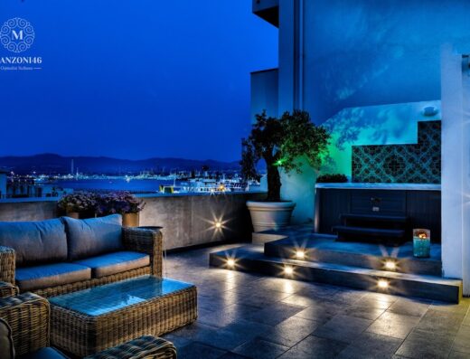 Terrazza panoramica di Manzoni 46, ospitalità siciliana, con divano in vimini, jacuzzi illuminata e vista notturna sulla città e sul porto.