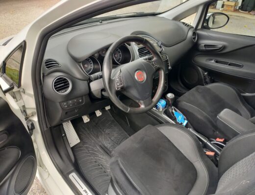 Interni di un'auto Fiat Punto bianca, con sedili sportivi neri e grigi, volante in pelle e pedaliera in alluminio.