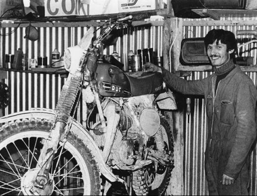 Immagine in bianco e nero di Mariotti Moto: un uomo sorridente appoggiato a una moto da cross d'epoca, probabilmente una Puch, in officina. Sullo sfondo scaffali con accessori e ricambi per moto.