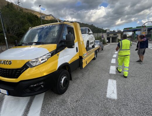 Marsicauto di Stefano Rino Soccorso Stradale: carro attrezzi Iveco giallo e bianco trasporta un'auto bianca su strada asfaltata, operatore in alta visibilità