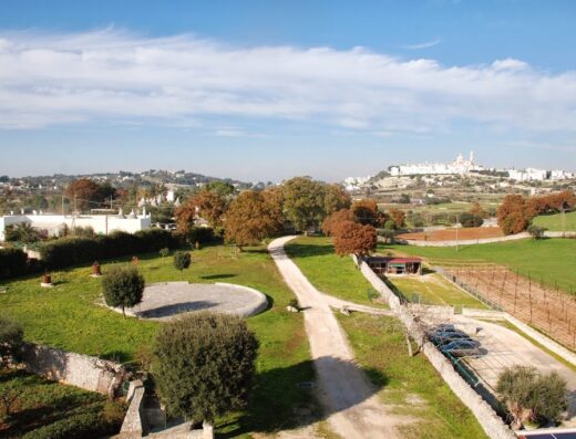 Veduta panoramica della Masseria Aprile, con strada sterrata, prati verdi, alberi e in lontananza il paese di Locorotondo, Puglia.