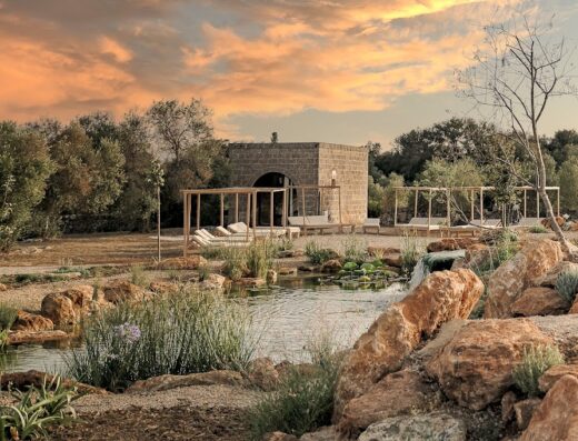Masseria Cuturi, Italy: Exterior view of the luxury masseria featuring a tranquil pond with lily pads, stone accents, lounge chairs under wooden canopies, and a picturesque sunset sky.