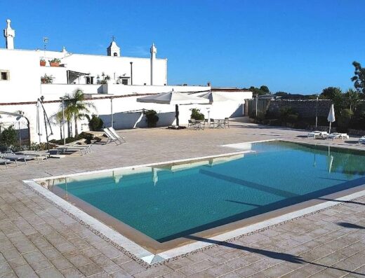 Masseria Del Crocifisso: ampia piscina con acqua cristallina, circondata da pavimentazione in pietra, lettini prendisole e ombrelloni bianchi, ideale per relax e vacanze in Puglia.