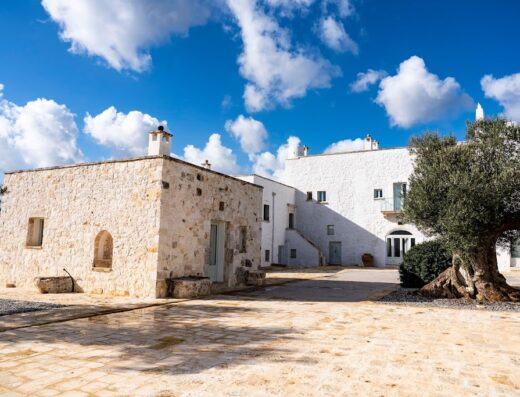 Masseria Fortificata Ayroldi: Veduta esterna della masseria con cielo azzurro e nuvole bianche, struttura in pietra e facciata bianca, ulivo secolare.