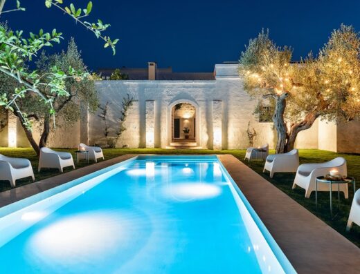 Masseria Narducci at night featuring a vibrant blue swimming pool, lounge chairs, olive trees with string lights, and a white stone building.