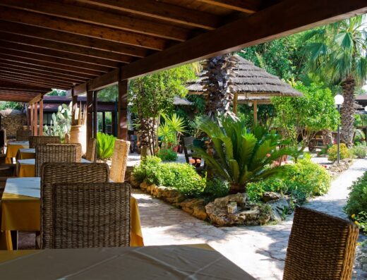 Ristorante all'aperto a Masseria Pisciani con tavoli apparecchiati, sedie in vimini, vegetazione lussureggiante e gazebo in paglia, per un'esperienza di viaggio indimenticabile.