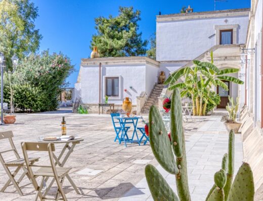 Masseria Prisuti: tranquillo cortile esterno con tavoli apparecchiati, cactus in primo piano e architettura tipica pugliese sullo sfondo, perfetta per un soggiorno rilassante.