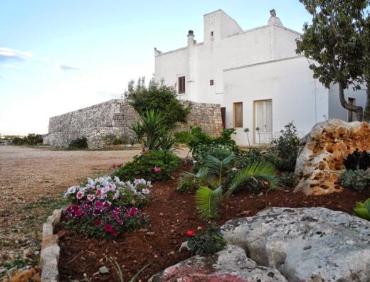 Image of Masseria Puglia Antica BB showcasing the building's exterior with a stone wall, white facade, garden with flowers, and natural rock formations.