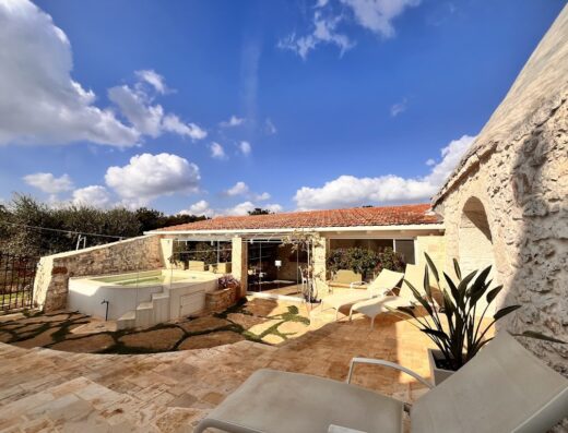 Vista esterna di trullo con piscina, lettini prendisole e cielo azzurro nuvoloso. Architettura tipica pugliese con materiali naturali.