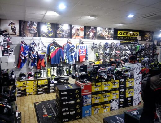 Interno del negozio di moto Mauri, con esposizione di abbigliamento tecnico da motocross, caschi Scorpion Exo e Airoh, stivali da moto e accessori. Poster di moto da cross sullo sfondo.