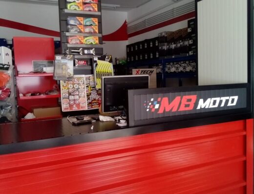 Interno del negozio MB Moto Shop con banco rosso, espositori di accessori moto, occhiali 100%, ricambi UFO Plast e insegna luminosa.
