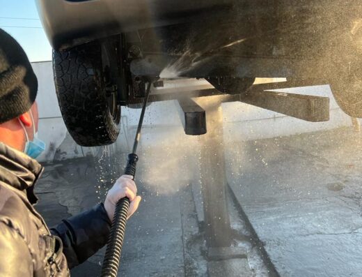 Un uomo lava il sottoscocca di un'auto con idropulitrice presso un autolavaggio, servizio di pulizia veicoli.