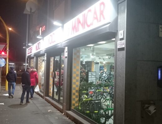 Foto della vetrina illuminata di Meddi & Sconci, negozio di moto e accessori a Roma. Persone camminano sul marciapiede di fronte al negozio.