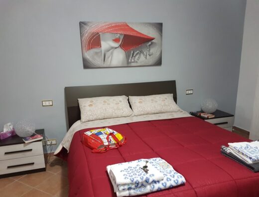 Camera da letto accogliente con letto matrimoniale, comodini, lampade, valigia rossa e quadro decorativo di una donna con cappello rosso.
