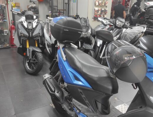 Esposizione di moto Miele in concessionaria: scooter blu con bauletto, moto grigia e altri modelli. Caschi e accessori visibili.