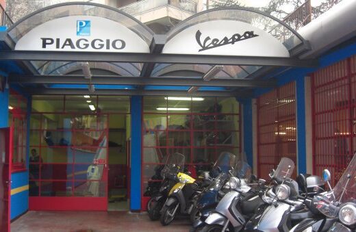Ingresso del centro assistenza autorizzato Piaggio e Vespa, con scooter parcheggiati davanti, insegne ben visibili. Servizi di riparazione e manutenzione per motocicli.