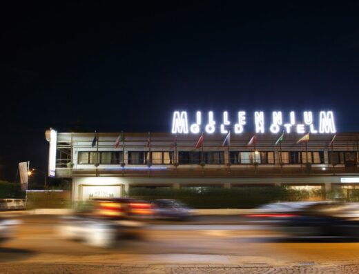 Esterno notturno del Millennium Gold Hotel illuminato, con bandiere e traffico in movimento, che offre servizi di ospitalità e alloggio di lusso.