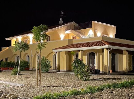 Minissale Farmhouse at night, a yellow building with a red tile roof is illuminated, showcasing its architectural details and landscaping. A car is parked nearby.