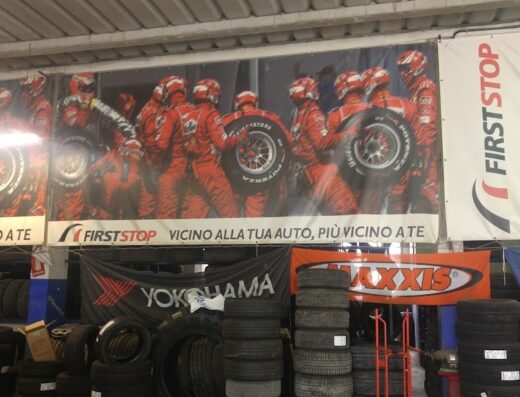 Interno di un negozio specializzato in pneumatici per auto e moto, con esposizione di gomme dei marchi Yokohama, Maxxis e Goodyear, e cartelloni pubblicitari di First Stop e team di Formula 1 con gomme.