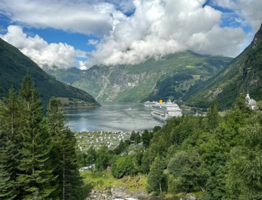 Vista panoramica mozzafiato sul Geirangerfjord in Norvegia con cascata in primo piano, montagne verdi, nuvole bianche e navi da crociera. Un'esperienza di viaggio indimenticabile.