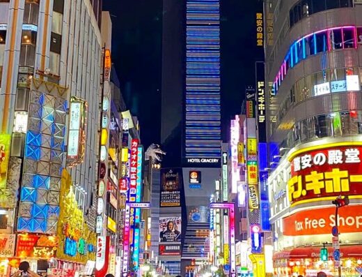 Affollata strada notturna a Tokyo, Giappone, illuminata da insegne al neon colorate e cartelloni pubblicitari. Il Hotel Gracery, con la testa di Godzilla, si staglia sullo sfondo.
