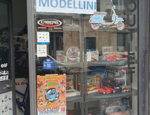 Vetrina di negozio a Ostuni con modellini di moto e auto, ricambi Piaggio, adesivo Vespa, poster Super Cartoonito Show e cartello Caberg Official Dealer. Accessori e marketplace per motociclisti.