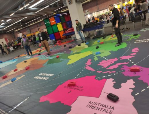 Grande mappa del mondo colorata a Modena Fiere con persone in piedi e piccoli carri armati giocattolo sparsi sopra, suggerendo un'attività o gioco interattivo.