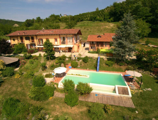 Veduta aerea di una casa di campagna con piscina nel Monferrato, circondata da colline verdi e vegetazione lussureggiante. Ideale per vacanze e relax.