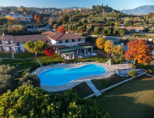 Vista aerea del Moniga Resort con piscina azzurra, prato verde curato, edifici con tetti rossi e panorama collinare sullo sfondo.