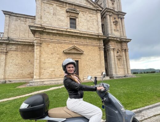 Una giovane donna sorridente indossa un casco e guida uno scooter grigio con bauletto davanti alla chiesa di San Biagio a Montepulciano, Toscana. Il servizio di noleggio scooter offre un'esperienza di viaggio unica alla scoperta della regione.