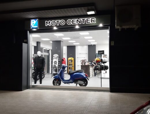Moto Center Snc: esterno del negozio con Vespa blu in vetrina, esposizione accessori moto e abbigliamento tecnico per motociclisti.